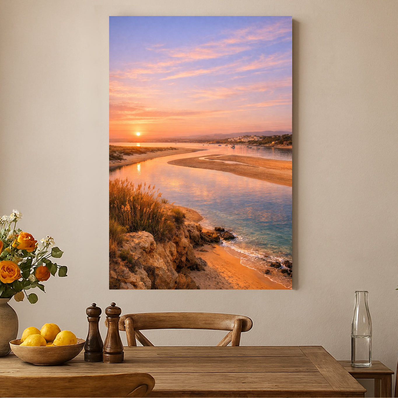 A vertical canvas wall art print depicting a coastal sunset scene with calm water, sandy shore, and a pink-orange sky, displayed above a wooden dining table in a styled interior.