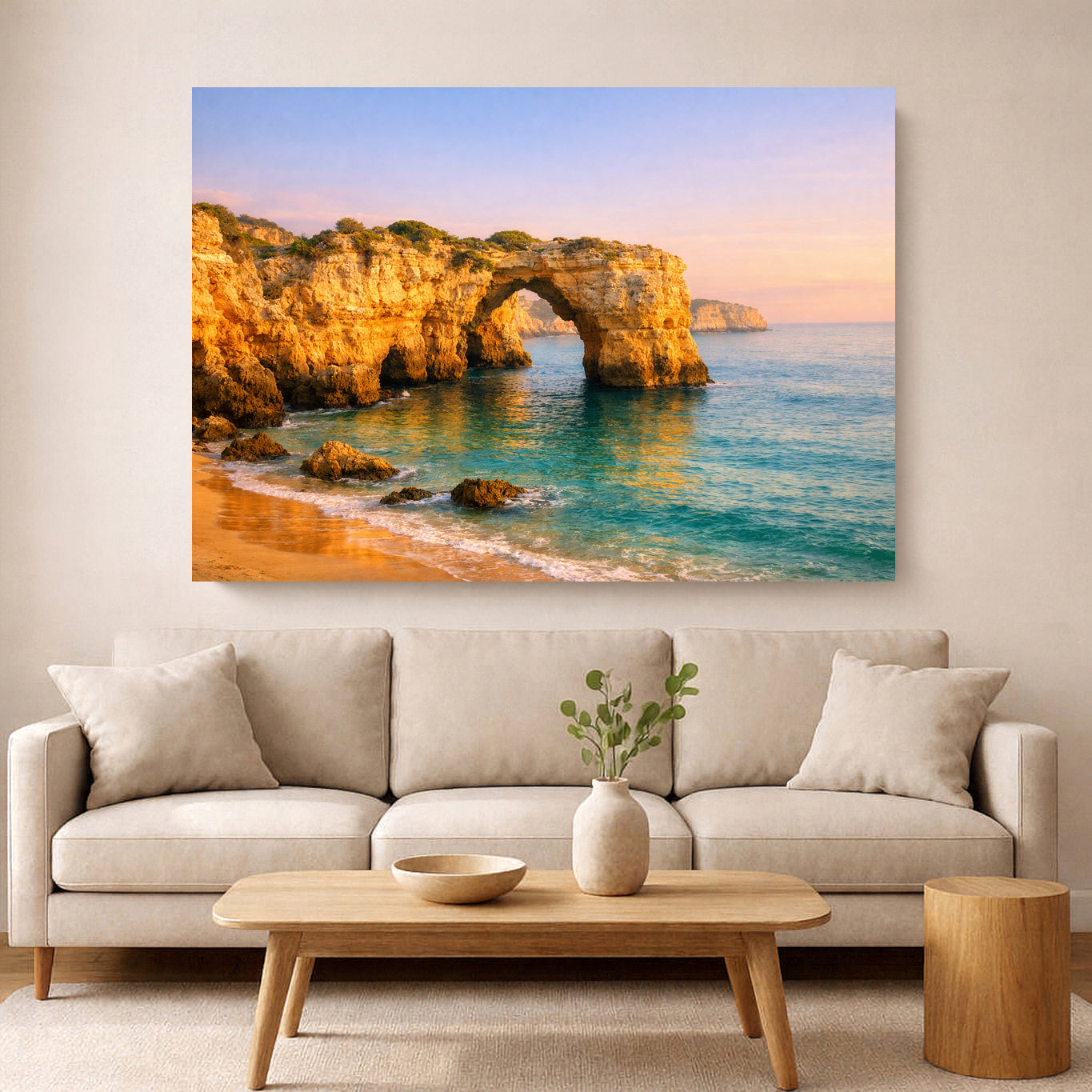 A large rectangular wall art print depicting a coastal rock arch and turquoise sea at sunset, displayed above a light beige sofa in a modern living room with a wooden coffee table and vase.