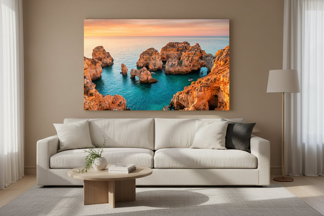 Sunset coastal scene with tall limestone cliffs, turquoise water, and two large sea stacks near the shore, with white cliff‑top buildings in the distance and small purple flowers in the rocky foreground.
