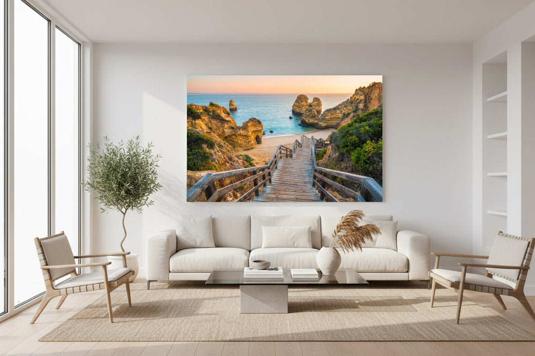 Large coastal landscape photograph of a wooden staircase leading down to a sandy beach between rocky cliffs at sunset, displayed above a white sofa in a modern minimalist living room.