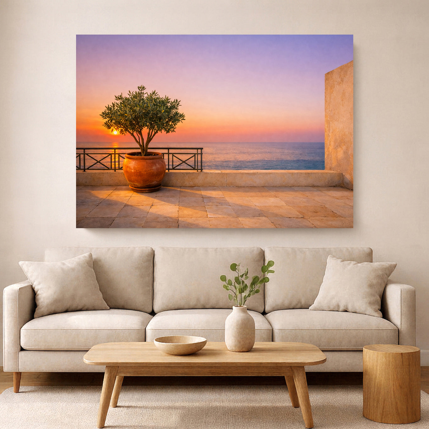A large rectangular wall art print showing a sunset seascape with a potted tree on a terrace, displayed above a light beige sofa in a modern living room.