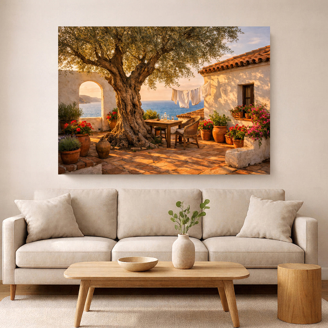 A large rectangular wall art print depicting a sunlit Mediterranean courtyard with a large tree, white buildings, potted flowers, and blue sea in the distance, displayed above a light beige sofa in a styled living room.